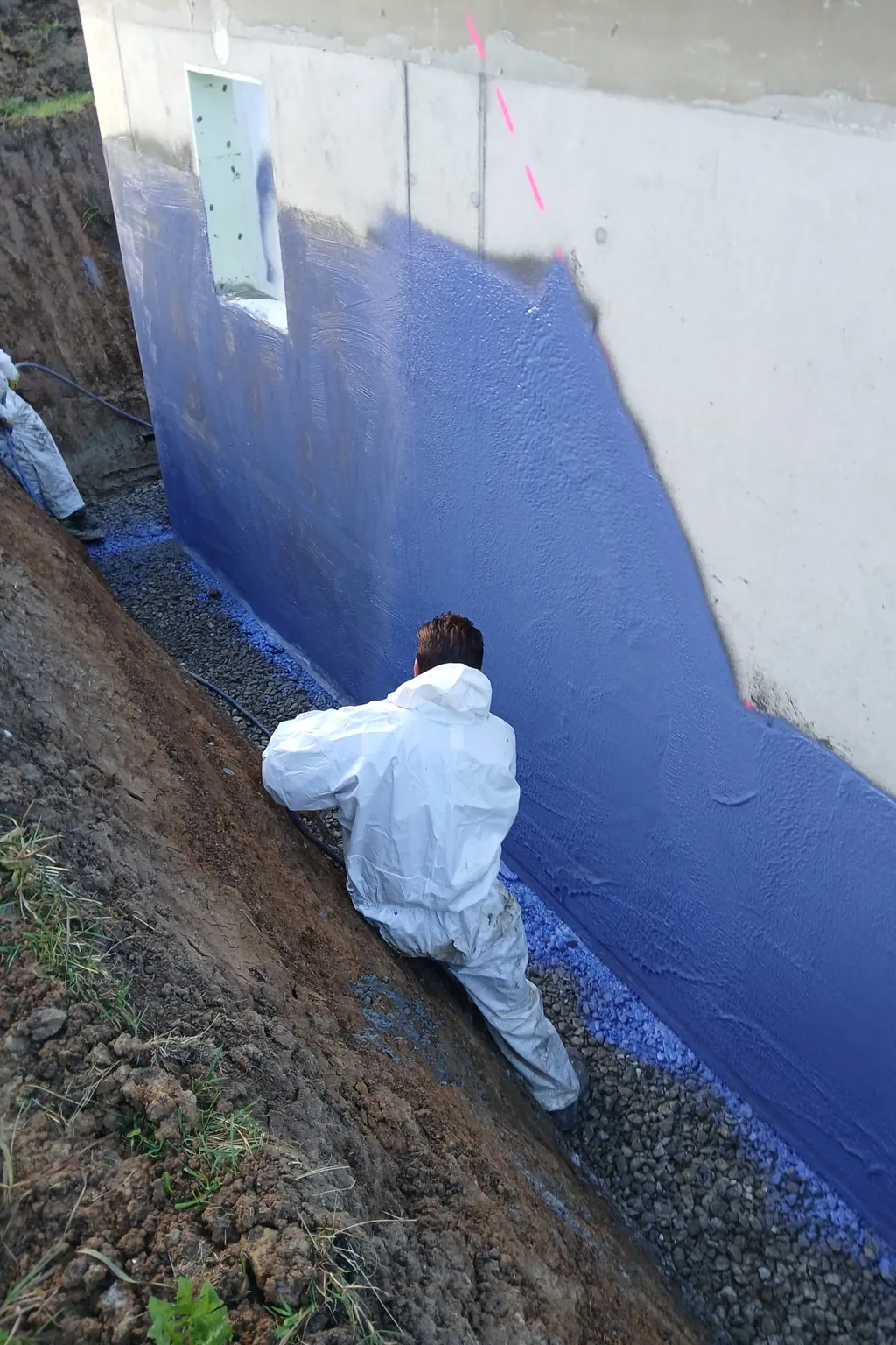 Zwei Mitarbeiter tragen die Abdichtung auf die Kelleraußenwand in der Baugrube auf.